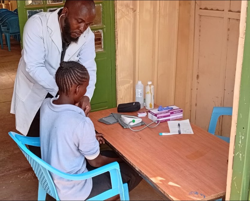A clinician in a white coat with a stethoscope examining a young patient seated in a blue chair beside a desk of medical supplies