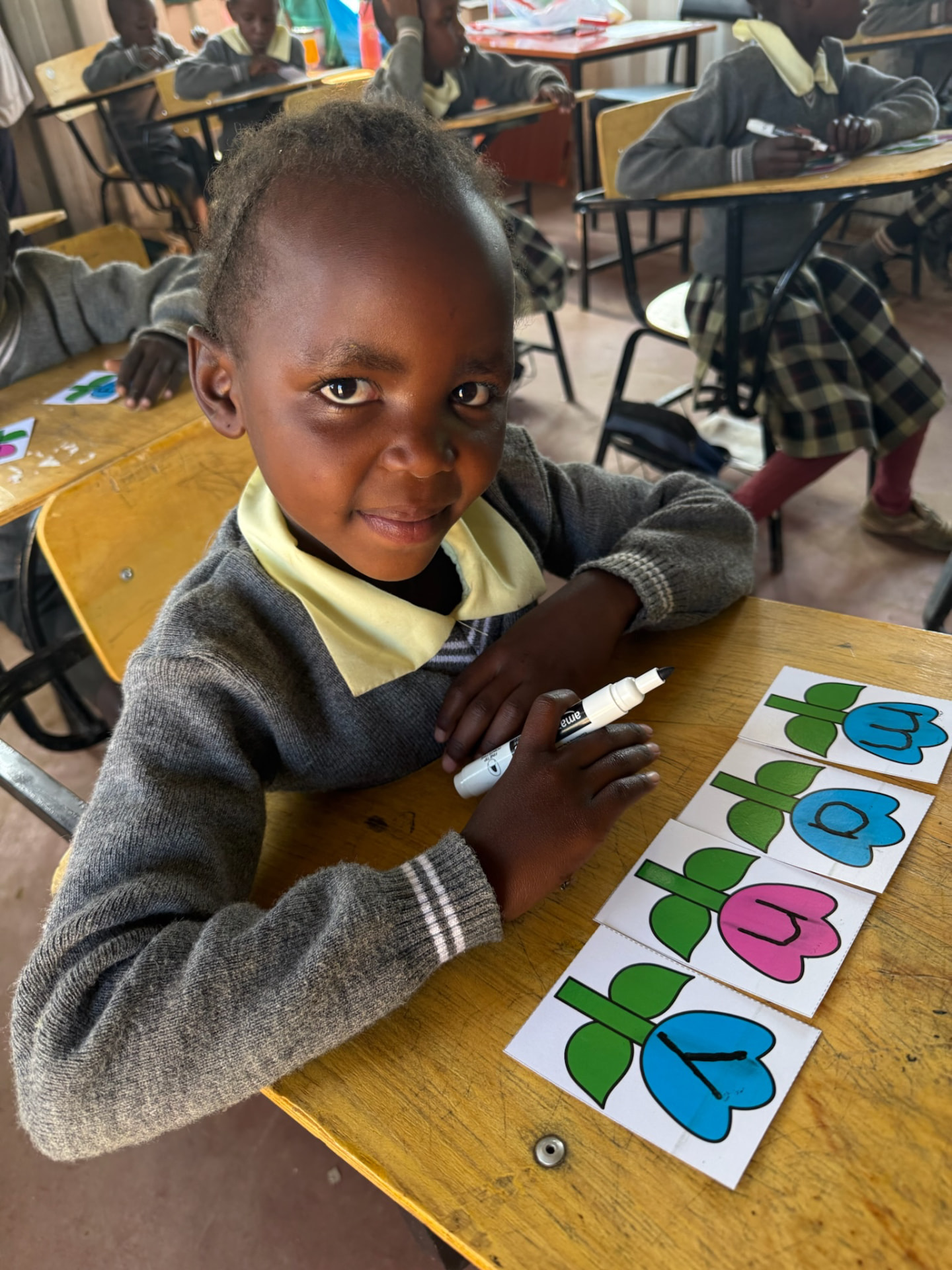 A young Kenyan student smiling at her desk, practicing letters with flower cards in her classroom